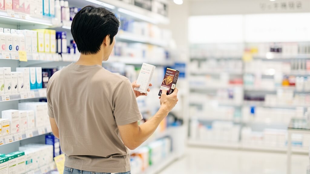 明るいドラッグストアの売り場で、2種類の発毛剤の箱を両手に持って見比べている30代日本人男性の後ろ姿の写真