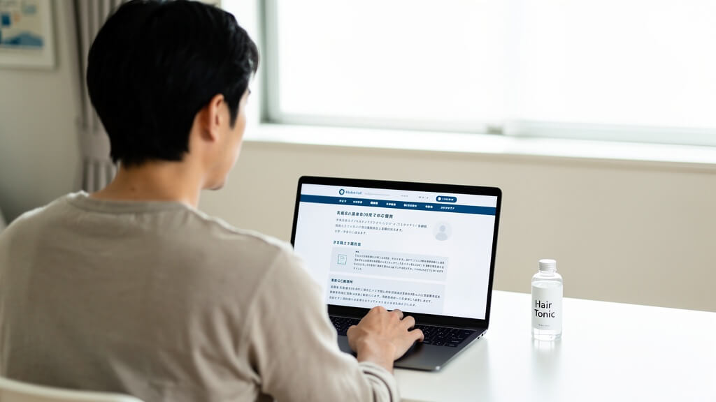 白いデスクに座った30代日本人男性がノートパソコンで医療相談サイトを見ながら次の薄毛治療の選択肢を調べている後ろ姿の写真