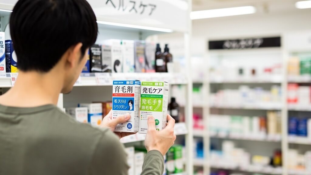 ドラッグストアの棚の前で、2つの発毛剤を見比べている30代日本人男性の後ろ姿。商品比較をしている場面を表した写真